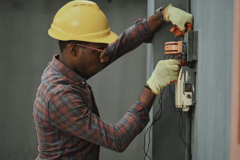 Technician in a yellow hard hat testing equipment on a wall-mounted panel